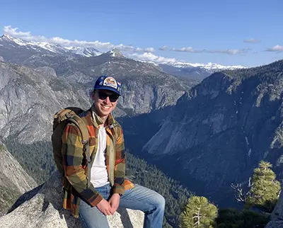 A person wearing a cap, sunglasses, and a plaid shirt sits on a rock with a mountainous landscape in the background. The sky is clear and the sun is shining, suggesting a pleasant day for hiking.