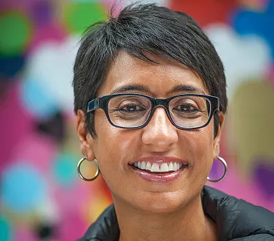 A person with short dark hair and glasses smiles against a colorful, abstract background. They wear hoop earrings and a dark jacket.