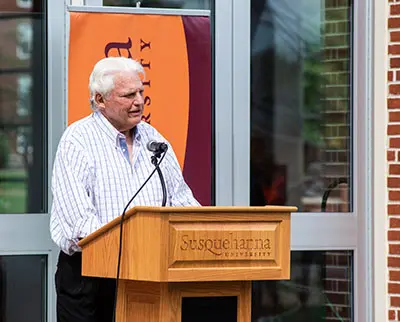 An elderly man with white hair speaks at a wooden podium with 