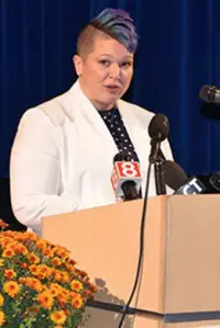 A person with short, colorful hair speaks at a podium adorned with microphones from various media outlets. They are wearing a white blazer and standing in front of a blue curtain with orange flowers on the side.
