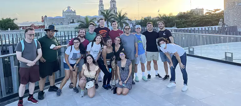 A group of people poses together on a rooftop with a cityscape in the background. The group includes both men and women, some standing and others crouching. The sun is setting, casting a warm glow over the scene.