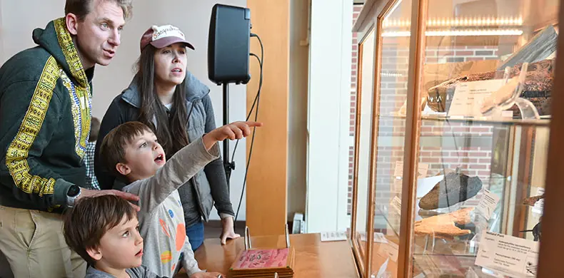A group of people, including two children and two adults, are looking at a museum display case. One child eagerly points at an exhibit inside the glass case, showcasing various objects. They appear engaged and curious.