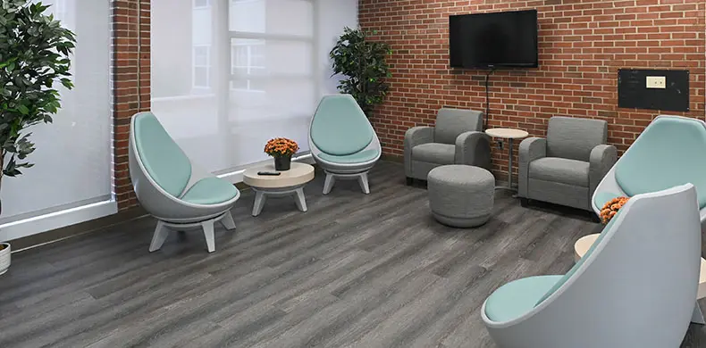 A modern waiting room with light blue and gray chairs, arranged in a circle. A round ottoman is in the center, surrounded by small tables with potted plants. A television is mounted on a brick wall. Large windows bring in natural light.