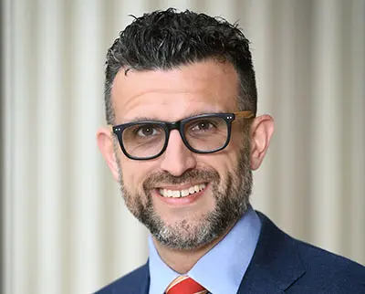 A man with short, curly hair and a beard wearing glasses, a blue shirt, a red tie, and a dark blazer is smiling at the camera. The background is softly blurred with vertical lines.