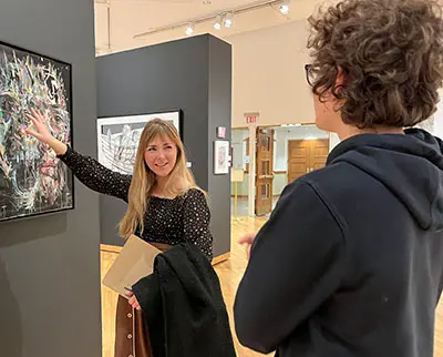 Two people are in an art gallery. A woman with long hair gestures at an abstract painting, smiling and holding a folder and coat. A person with curly hair and glasses listens, partially turned away from the camera.