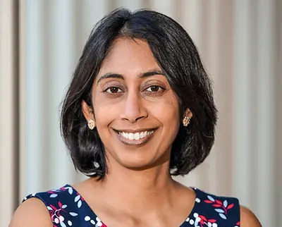 A person with shoulder-length black hair smiles at the camera. They are wearing a floral-patterned dress and gold earrings. The background is softly blurred vertical lines.