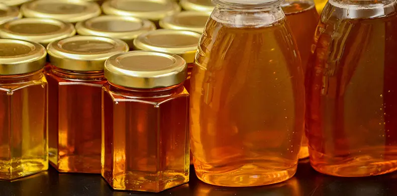 Rows of glass jars and plastic bottles filled with golden honey, each sealed with a golden lid, are arranged closely together on a dark surface, reflecting light and showcasing the amber liquid.
