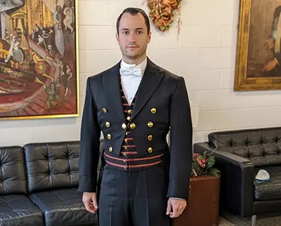 A man stands indoors wearing a formal black tailcoat with gold buttons, a white bow tie, and a striped waistcoat. He is in a room with leather sofas and framed art on the walls.