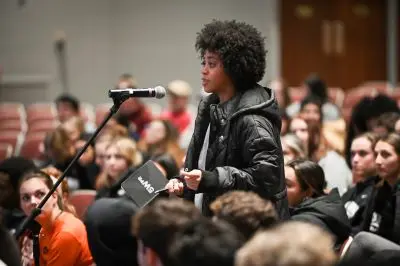 A person with curly hair speaks into a microphone while standing in an auditorium filled with people. They hold a notebook and wear a black jacket, appearing engaged in a discussion or presentation.