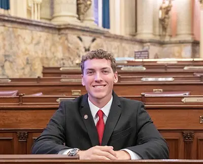 A person in a suit and red tie sits smiling in a formal setting with wooden desks and ornate walls.