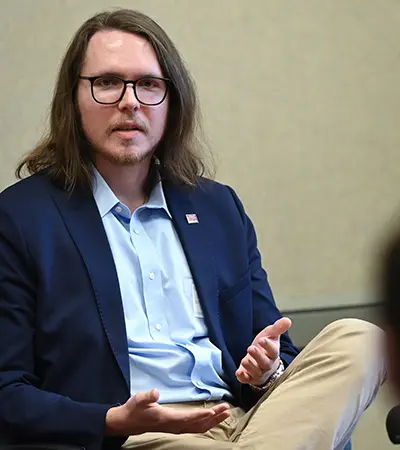 A man with long hair and glasses, wearing a blazer and blue shirt, gestures with one hand during a discussion. He is seated, with a neutral background.