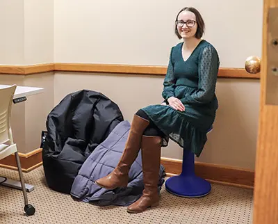 A person in a green dress and brown boots sits on a blue stool in a corner of a room. There is a black bean bag and a gray blanket on the floor nearby. The room has beige walls and a wooden door.