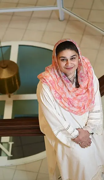 A person wearing a white outfit and a floral pink scarf stands smiling on a staircase. The floor is tiled, and part of a circular design is visible below. A decorative hanging object is in the background.