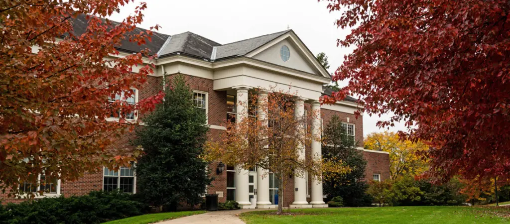 A large brick building with white columns is surrounded by autumn trees with red and yellow leaves. A pathway leads to the entrance, and the overcast sky complements the fall setting.