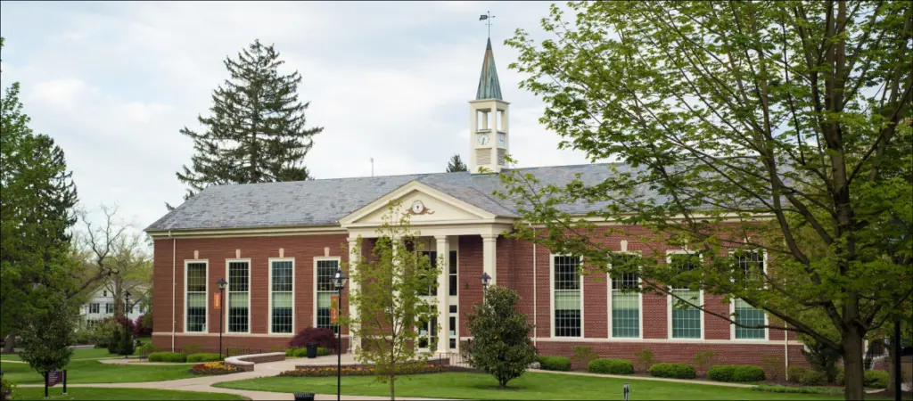 A red brick building with large windows and a white clock tower sits surrounded by lush green trees and grass. The sky is cloudy, and a winding path leads to the entrance.