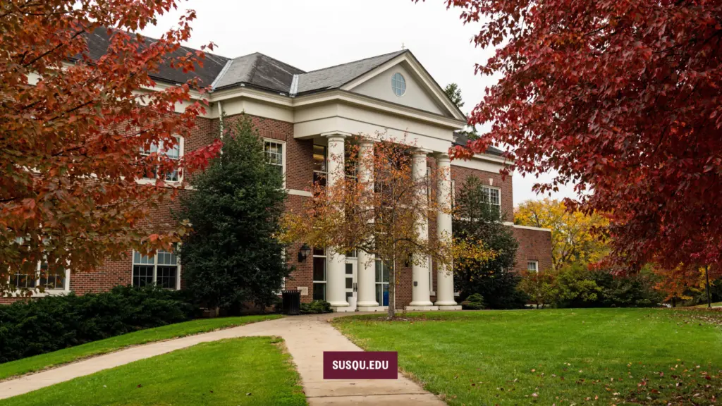 A large brick building with white columns is surrounded by green grass and trees with red and yellow autumn leaves. A pathway leads to the entrance. A sign reading 