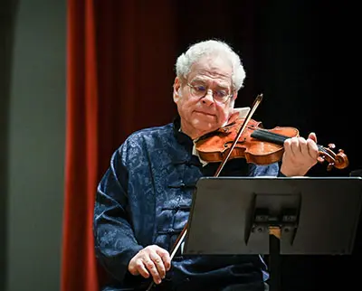 An elderly musician with glasses plays the violin while reading sheet music on a stand, set against a dark and red curtain backdrop.