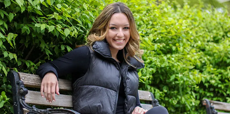 A person with long hair sits on a wooden park bench, smiling, wearing a black puffer jacket and dark pants. Green bushes are visible in the background.