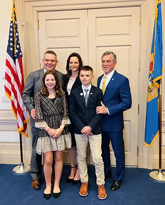 A group of five people posing in a room with an American flag and another flag in the background. Three adults stand behind two children. They are all dressed formally and smiling.