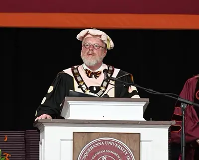 A person in academic regalia, including a cap and gown, stands at a podium with a microphone, delivering a speech. The podium features a logo for 体育买球 University, and a maroon and orange backdrop is visible.