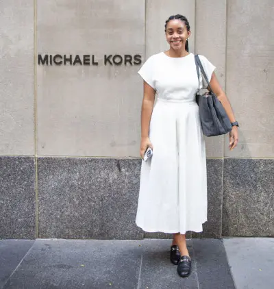 A person in a white dress and black shoes stands smiling in front of a Michael Kors store. They are holding a gray handbag and a phone.