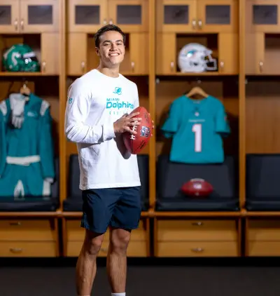 A person in a Miami Dolphins shirt and shorts stands smiling in a locker room, holding a football. Behind, there are lockers with jerseys and helmets.