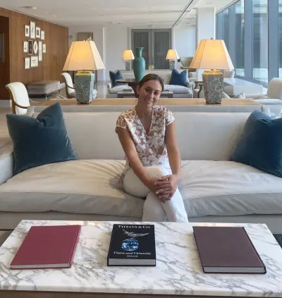 A person sits on a white sofa in a modern, well-lit living room with large windows. The room features decorative lamps and cushions. A marble coffee table in front holds three books, including one titled 