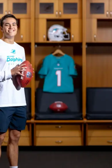 A man is standing in a sports locker room holding a football. He is wearing a white long-sleeve shirt and dark shorts. Behind him are lockers with sports memorabilia, including a teal jacket, a jersey, and tennis rackets.