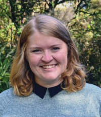 Smiling person with shoulder-length hair, wearing a gray sweater with a dark collar, stands in a wooded outdoor setting.