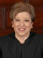 A person with short, light brown hair is smiling at the camera. They are wearing pearl earrings and a black judicial robe. The background features wooden paneling.