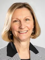A person with short, light-colored hair is smiling. They are wearing a black and white houndstooth blazer and earrings. The background is plain and light-colored.