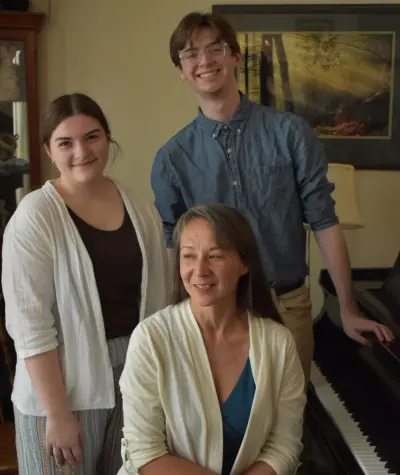 A group of three people indoors, with two standing behind a seated woman. They are gathered near a piano, all smiling. The room has a peaceful ambiance with a framed nature picture on the wall.