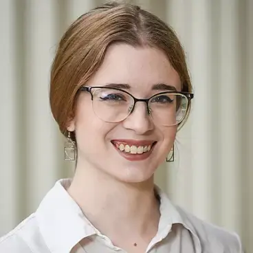 A person with long hair, wearing glasses and square earrings, smiles while looking at the camera. They are dressed in a light-colored, button-up shirt against a muted background.
