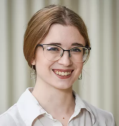 A person with long hair, wearing glasses and square earrings, smiles while looking at the camera. They are dressed in a light-colored, button-up shirt against a muted background.