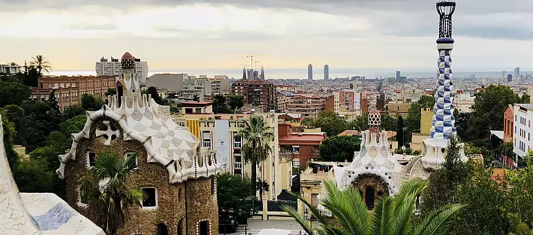 A panoramic view of Barcelona from Park Güell, featuring colorful, mosaic-covered buildings. The skyline has several tall buildings, and the Sagrada Familia is in the distance under a cloudy sky. Lush greenery surrounds the area.