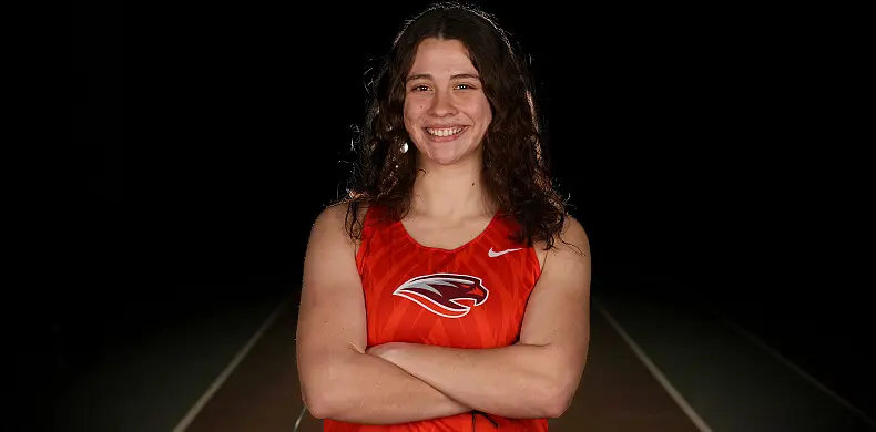 A woman stands confidently with her arms crossed, wearing an orange sports jersey with a panther logo. She has long, curly hair and is smiling in front of a dark background.