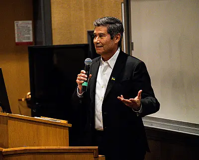 A man in a suit holds a microphone, standing at a wooden podium. He appears to be giving a presentation or speech in a room with a whiteboard and a monitor in the background.