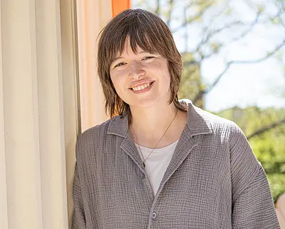 A person with short hair is smiling while leaning against a light-colored pillar. They are wearing a textured, button-up shirt. The background features a blurred outdoor setting with trees and a clear sky.