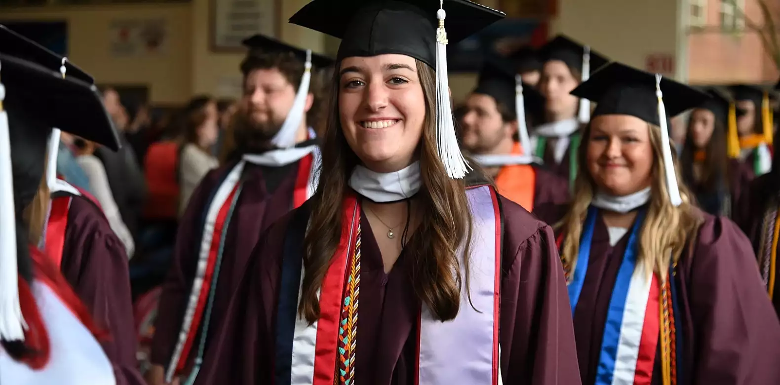 A group of graduates in maroon caps and gowns stand indoors, with a young woman in the foreground smiling. She is wearing honor cords and a white stole with red accents. Other graduates are visible in the background.
