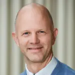 A bald man wearing a blue sweater over a white collared shirt smiles at the camera. The background is a soft, striped pattern in muted colors.