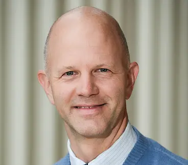 A bald man wearing a blue sweater over a white collared shirt smiles at the camera. The background is a soft, striped pattern in muted colors.