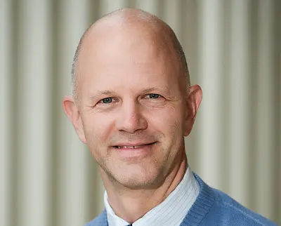 A bald man wearing a blue sweater over a white collared shirt smiles at the camera. The background is a soft, striped pattern in muted colors.
