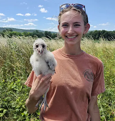 A person smiling while holding a fluffy young owl in an open field. They are wearing a peach-colored t-shirt and sunglasses on their head. The background features grass and a clear blue sky with a few clouds.