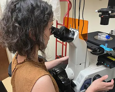 A person with curly hair in a brown top looks into a microscope in a lab setting, focusing on specimen slides. The microscope is equipped with various attachments and there are lab items in the background.