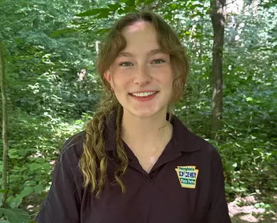 A person with a ponytail wearing a black shirt stands in a lush green forest. They are smiling at the camera, and the shirt has a logo with text. Sunlight filters through the trees, creating a serene atmosphere.