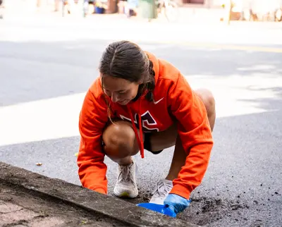 A person in an orange hoodie crouches by a roadside, wearing gloves and working with soil or debris. They appear focused on a task, possibly related to cleaning or gardening, on a sunny day.