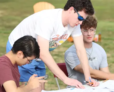 Three people are gathered around a table outdoors, engaging in an activity. One person is writing, another is looking at a phone, and the third is pointing at a document. They are dressed casually, and the background is grassy with chairs.