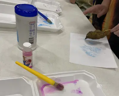 Hands painting on paper with brushes and paint. There are paintbrushes, a pink paint bottle, and a container on the table. A brown object on a stick is used for stamping blue paint onto the paper.