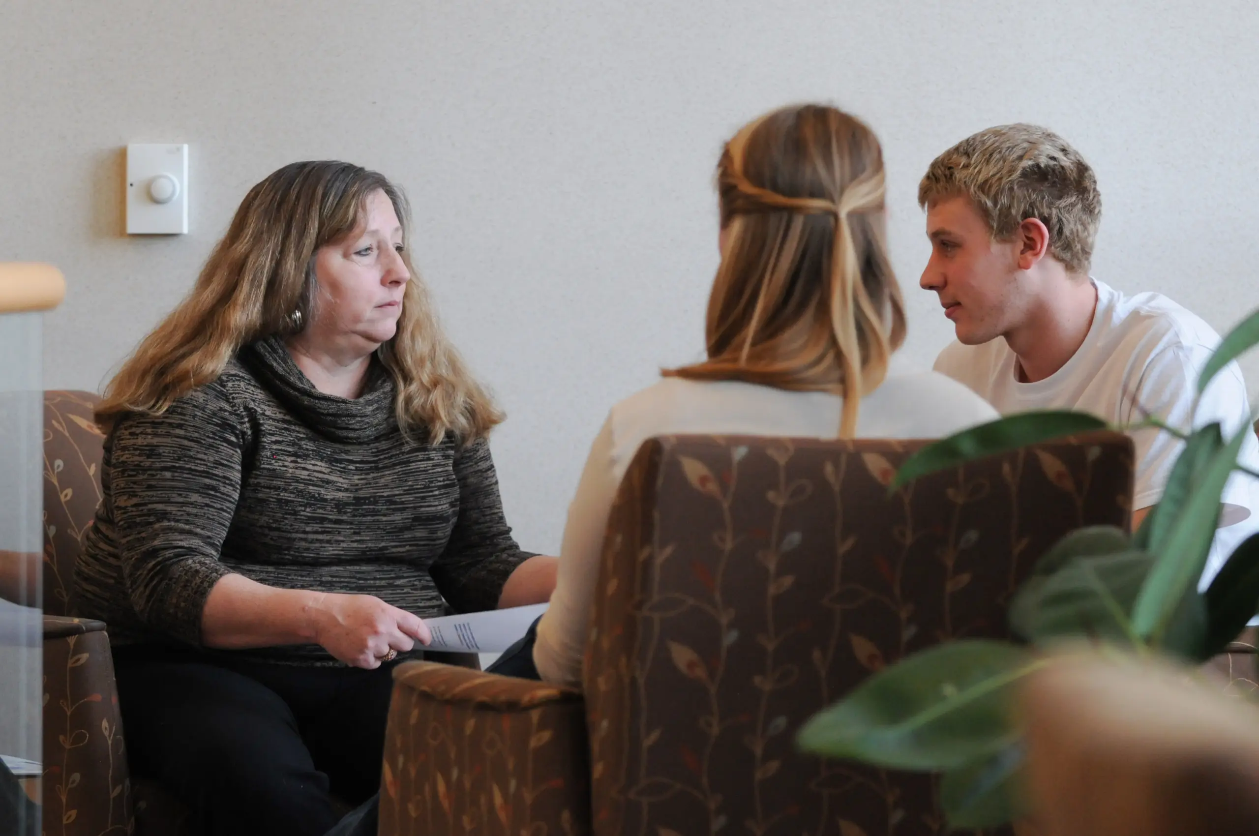 Three people are engaged in a conversation while seated in cushioned chairs. A woman with long hair listens intently to a young man and a woman seated across from her. The setting appears to be informal, with a plant visible in the foreground.