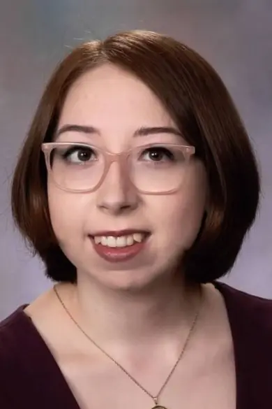 A person with shoulder-length brown hair and glasses is smiling. They are wearing a dark purple top and a necklace. The background is a soft gradient of gray and blue tones.